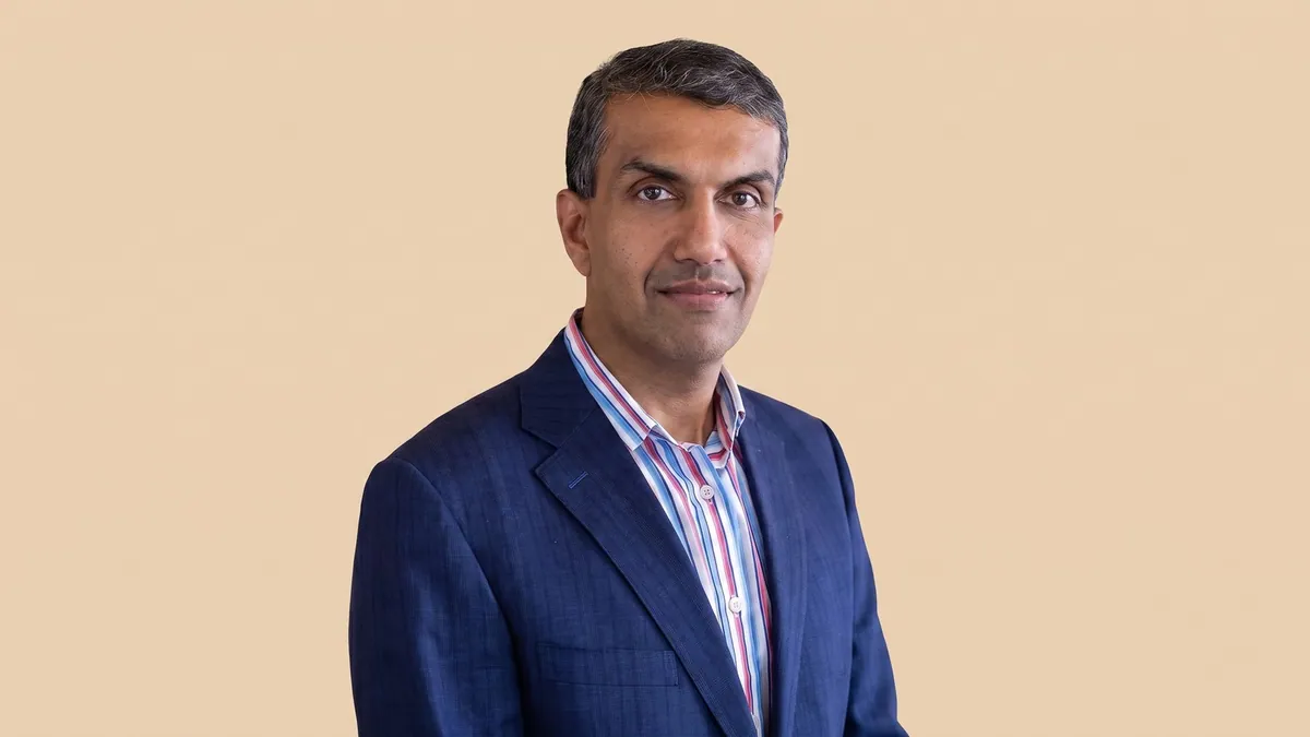 Portrait of Anil Chakravarthy in a navy suit and striped shirt against a flat pastel warm sand backdrop