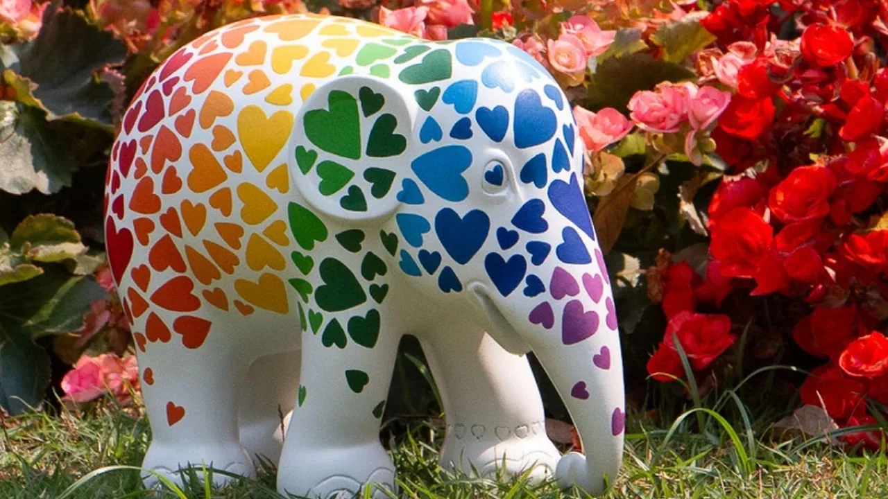 A white elephant sculpture painted with rainbow hearts, standing in red flowers, in Elephant Parade's launch image