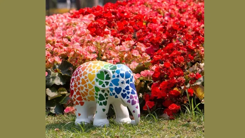 A white elephant sculpture painted with rainbow hearts, standing in red flowers, in Elephant Parade's launch image