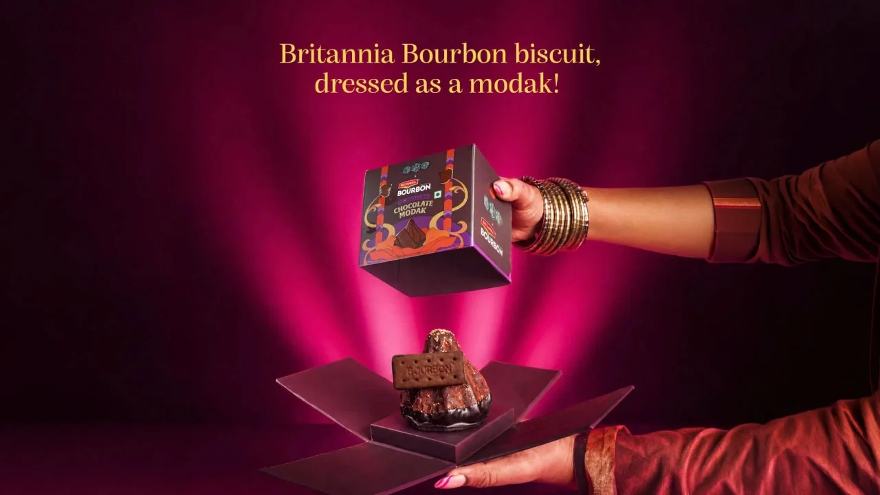 Hands lifting a box off a Bourbon Chocolate Modak on a magenta set, in Britannia Bourbon and Bombay Sweet Shop's key visual