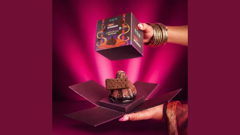 Hands lifting a box off a Bourbon Chocolate Modak on a magenta set, in Britannia Bourbon and Bombay Sweet Shop's key visual