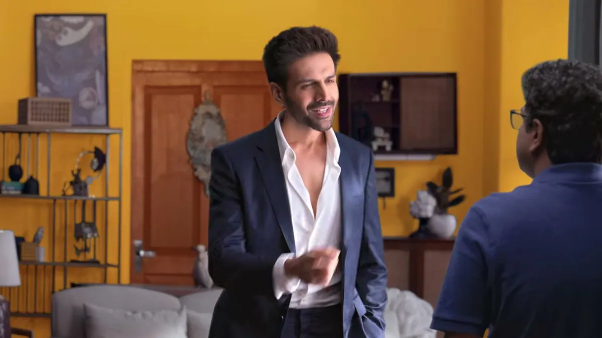 Kartik Aaryan as the Macaw Genie in a navy suit, in a freshly painted yellow living room, from the Macaw Paints festive film