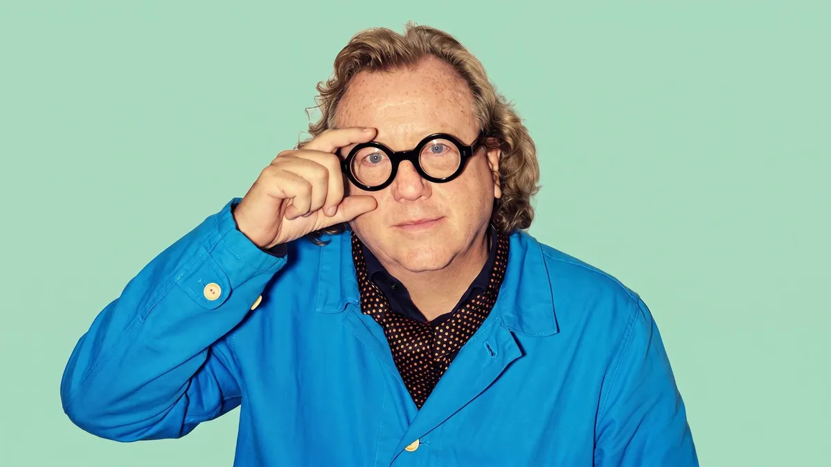 Portrait of Simon Shaw in a blue jacket holding round spectacles to his eye, against a flat pastel mint backdrop