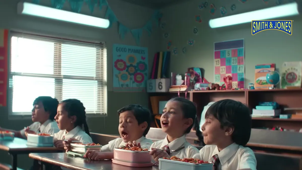 Five schoolchildren in uniform eating pasta from tiffin boxes at their classroom desks, from the Smith & Jones film