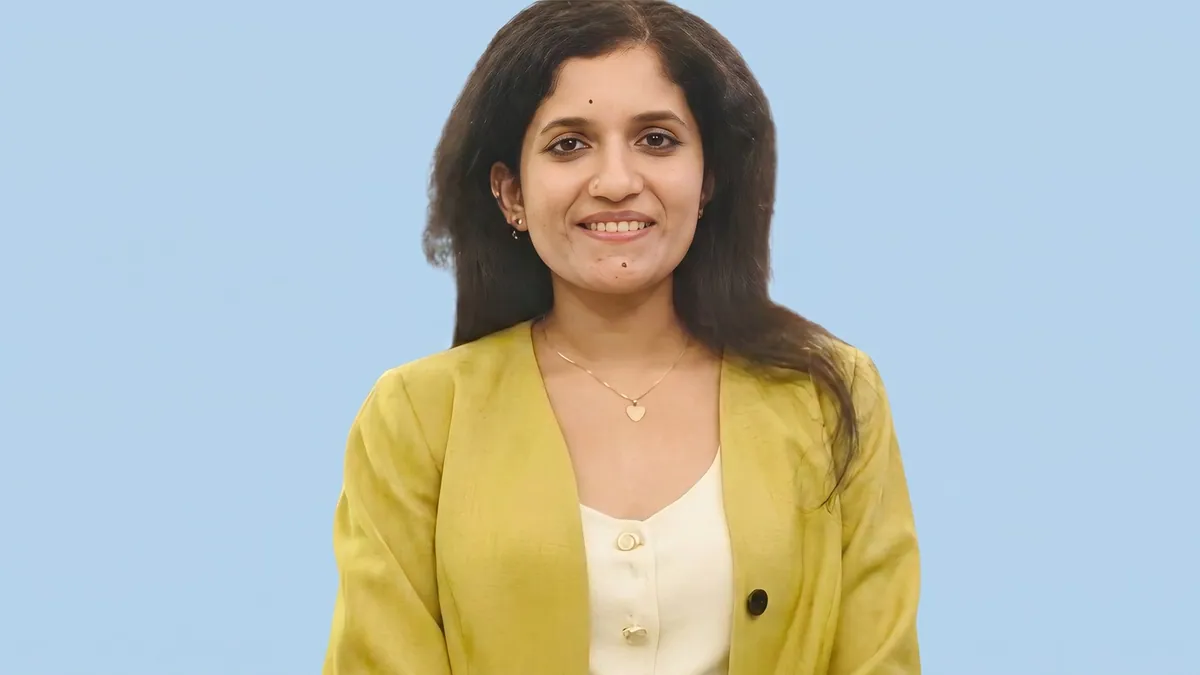Portrait of Sugatha Karthikeyan in an olive jacket against a flat pastel powder blue backdrop