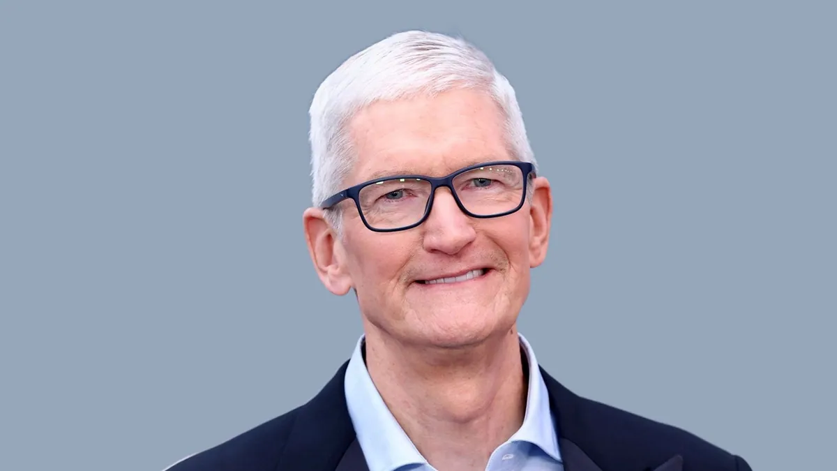 Tim Cook in a navy blazer and pale blue open-collar shirt, wearing dark rectangular glasses